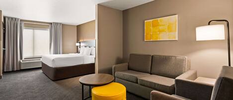 Une chambre d’hôtel avec un lit, un canapé, un pouf jaune, une petite table et une lampe.