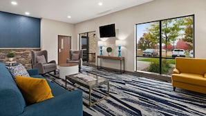 A modern hotel lobby with a blue and white striped carpet, a large window with a view of the outside, a flat-screen TV mounted on the wall, and a seating area with a blue sofa and a yellow armchair. - Comfort Suites Hudson I-94 (Hudson)