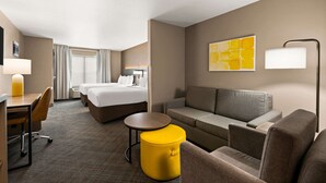 A hotel room with a bed, a sofa, a small table, a lamp, and a yellow ottoman. - Comfort Suites Hudson I-94 (Hudson)
