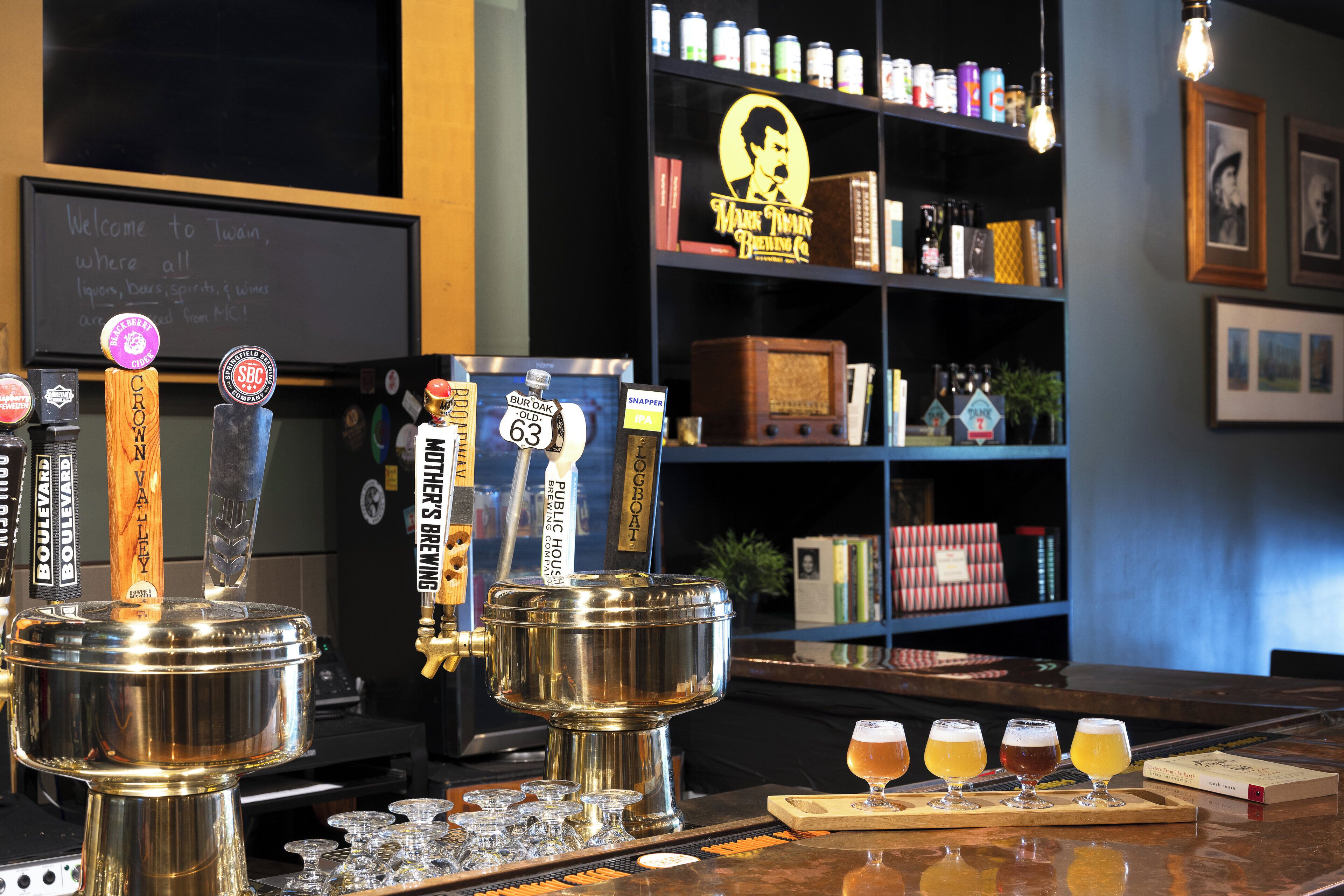 A bar area with tap handles labeled with beer names, a selection of beers on a tray, and a bookshelf in the background.