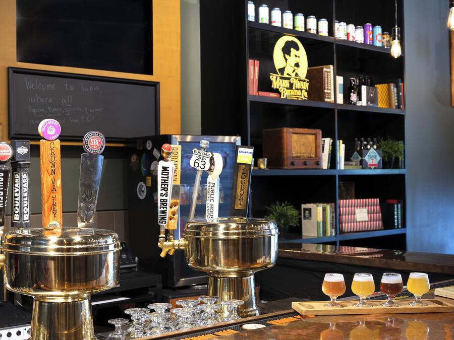 A bar area with tap handles labeled with beer names, a selection of beers on a tray, and a bookshelf in the background.