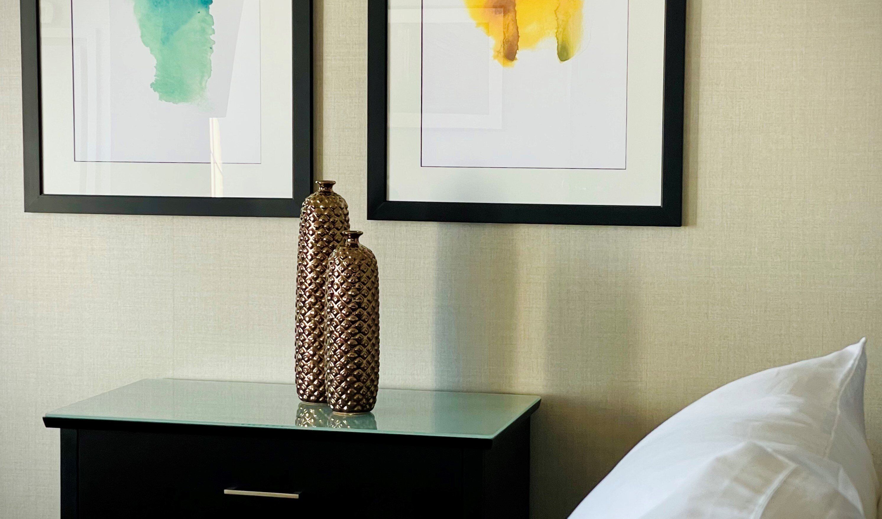 A hotel room with a bed, a nightstand with a decorative bottle, and two framed abstract paintings on the wall.