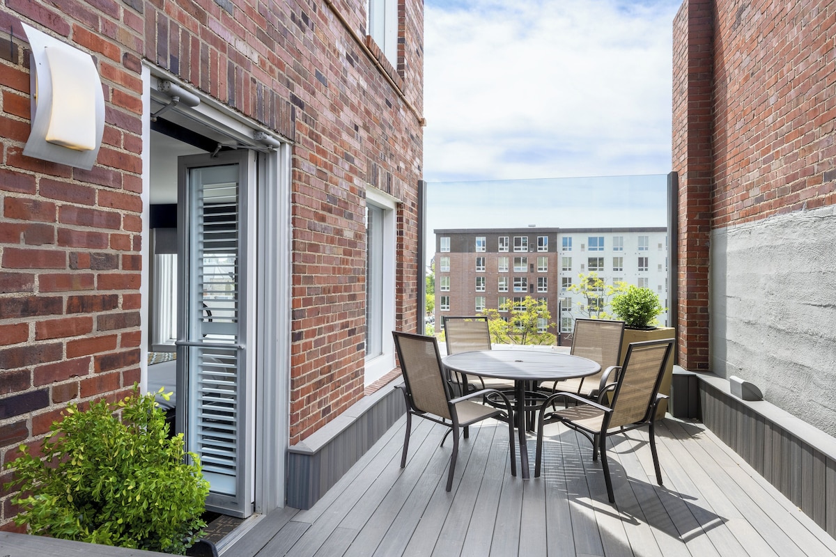 A wooden deck with a round table and chairs, a small potted plant, and a brick building in the background.