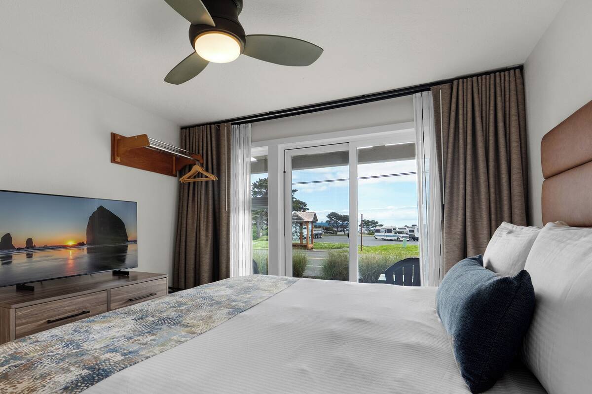 A hotel room with a bed, a television, a ceiling fan, and a view of the outdoors.