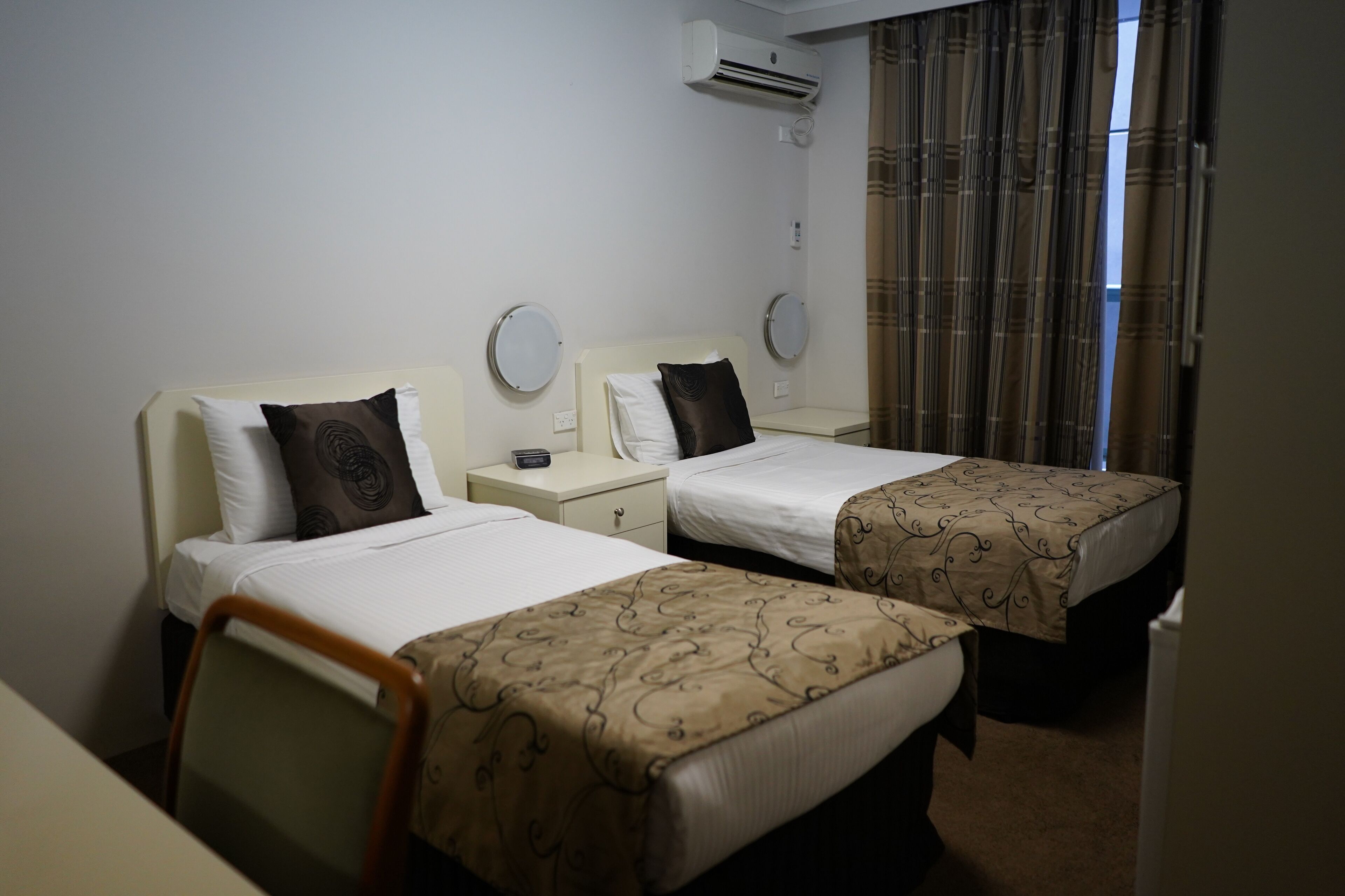 Ein Hotelzimmer mit zwei Einzelbetten, jeweils mit Nachttisch und Spiegel. Es gibt ein Fenster mit Vorhängen, eine Klimaanlage an der Wand und einen Sessel in der Ecke.
