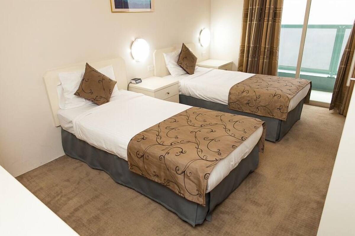 Quarto de hotel com duas camas, cada uma com travesseiro e mesa de cabeceira. Há arandelas acima das camas e uma janela com cortinas.