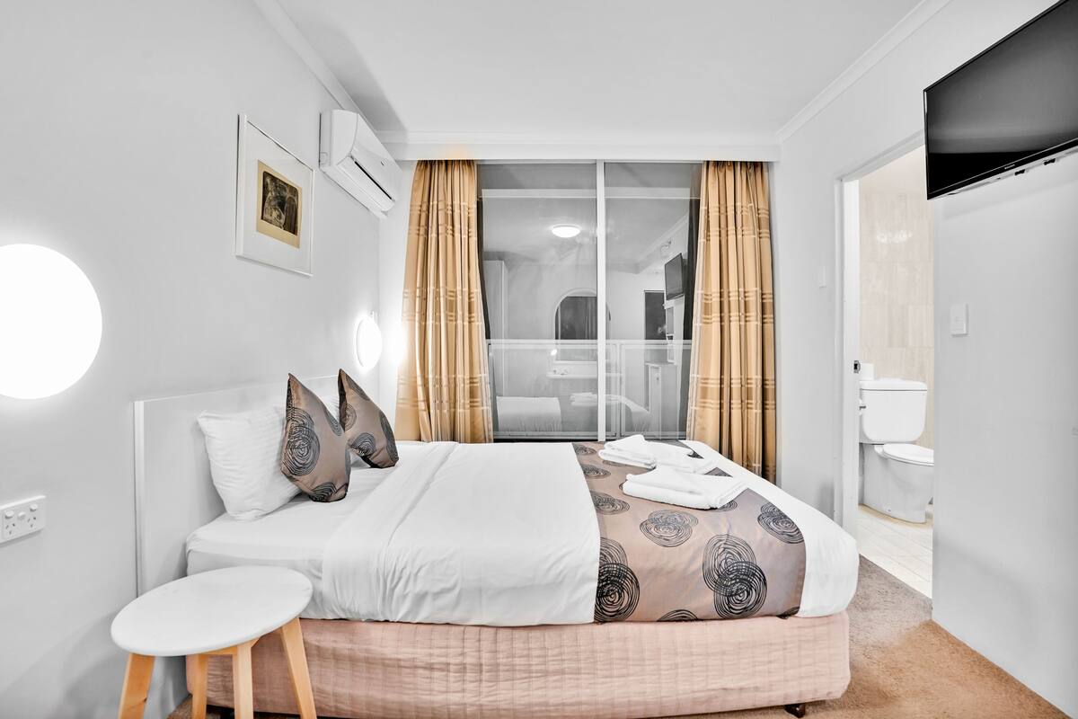 Quarto de hotel moderno com uma cama grande, um banquinho pequeno, uma TV e um banheiro visível através de uma porta de vidro.