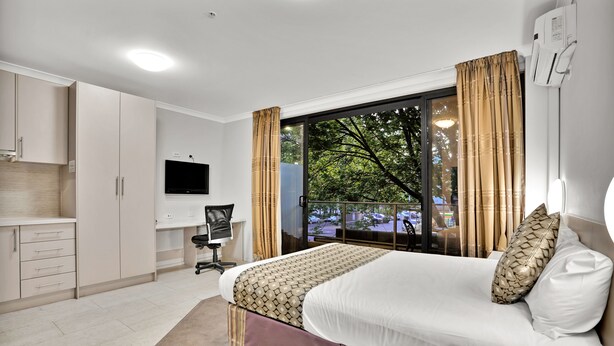 A hotel room with a bed, a desk, a chair, a TV, and a balcony with a view of trees and a parking lot. - Carlton Lygon Lodge (Carlton)