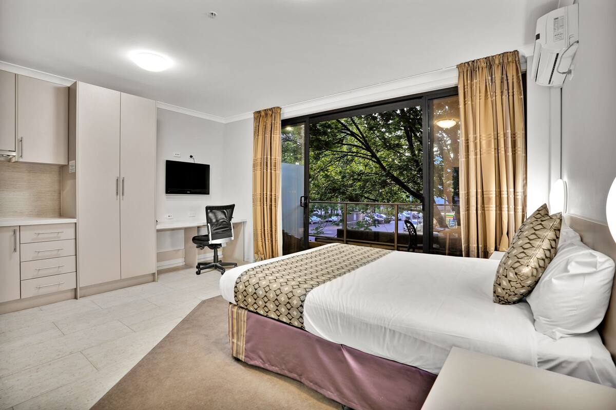 Quarto de hotel com cama, escrivaninha, cadeira, televisão e varanda com vista para árvores e estacionamento.