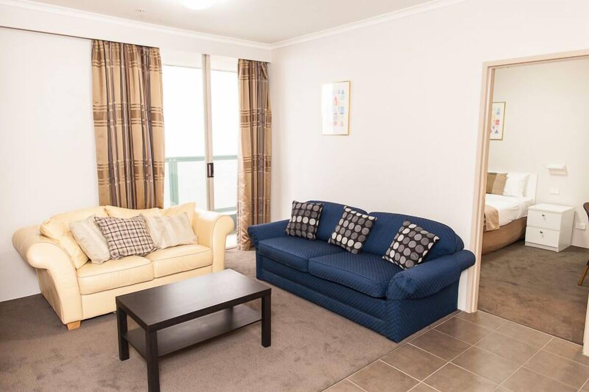 Sala de estar com um sofá bege, um sofá azul e uma mesa de centro. Há uma porta que dá para um quarto com uma cama e uma mesinha de cabeceira.