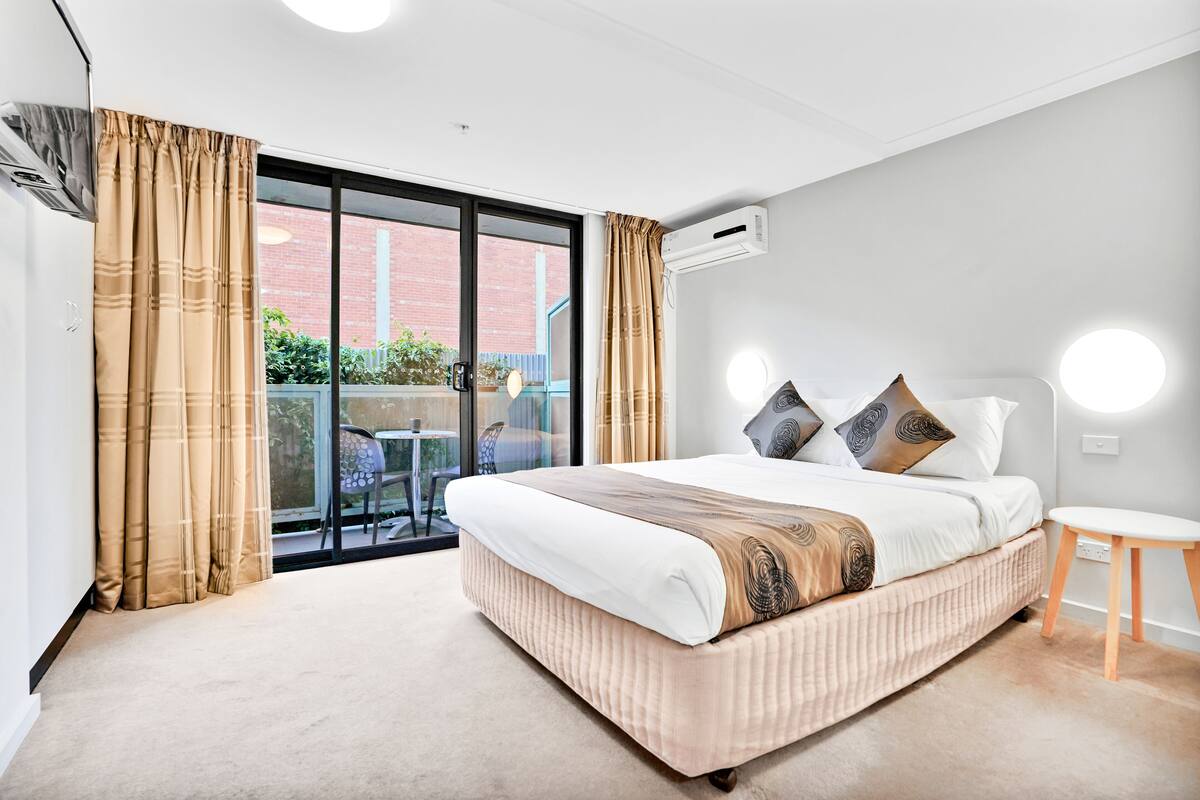 Quarto moderno com uma cama grande, uma mesinha e vista para um edifício de tijolos através de uma porta de correr de vidro.