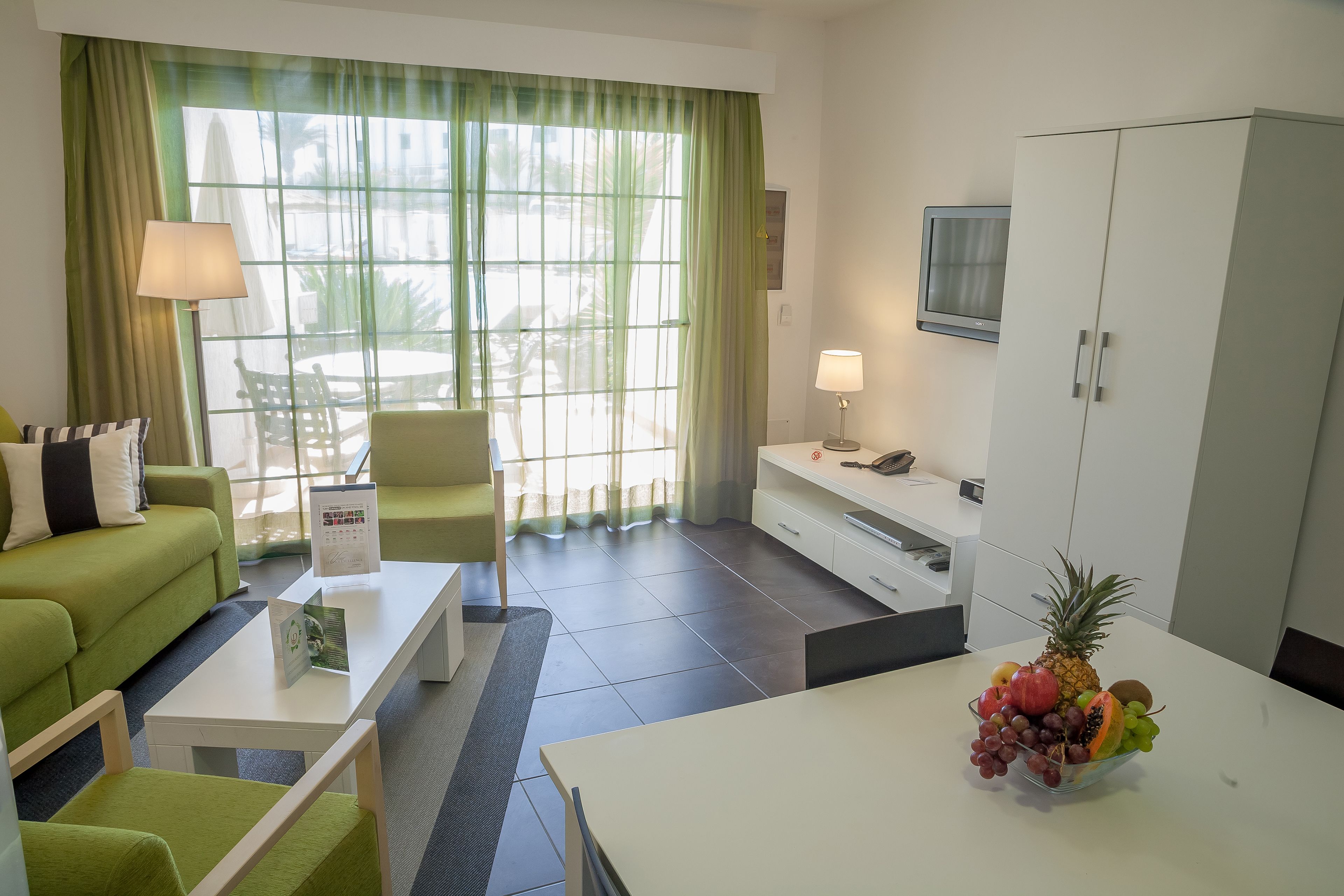 A modern hotel room with a green sofa, a white coffee table, a dining table with a fruit bowl, and a TV on the wall.