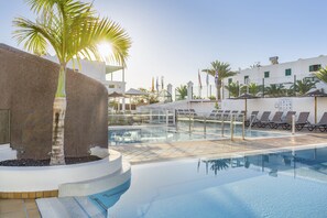 A pool area with lounge chairs, a palm tree, and a building with flags. - Club Del Carmen (Tías)