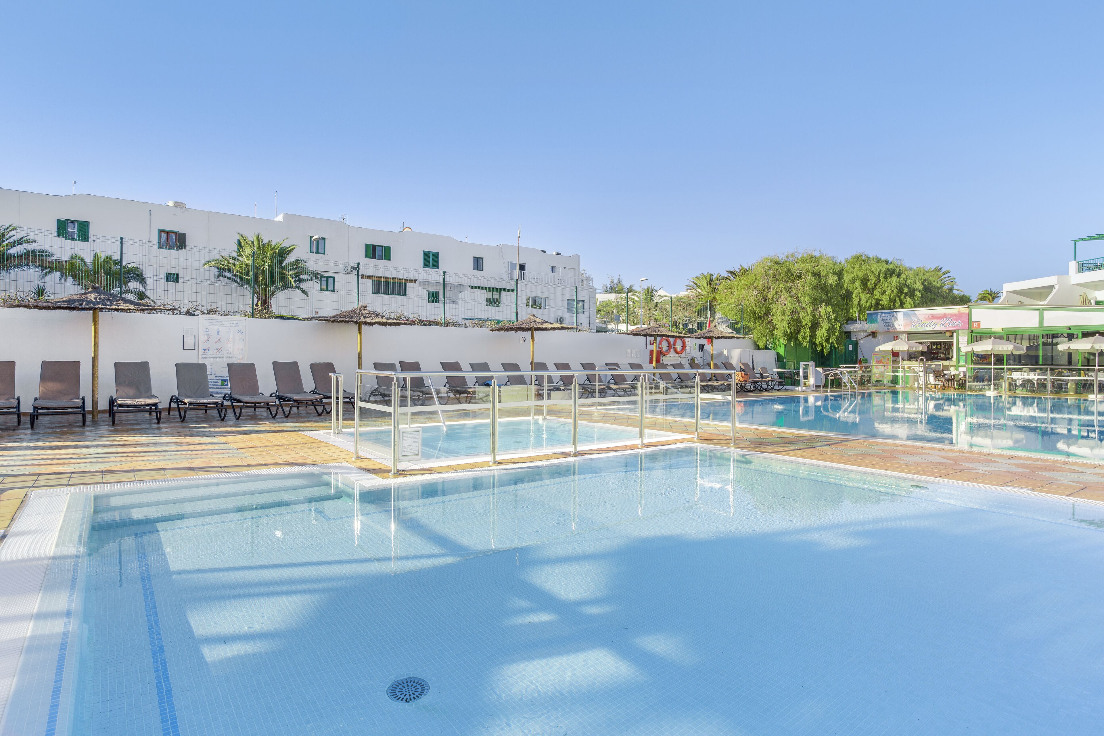 A swimming pool with lounge chairs and umbrellas, a white building with multiple windows, and palm trees in the background.
