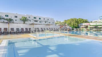 A swimming pool with lounge chairs and umbrellas, a white building with multiple windows, and palm trees in the background.