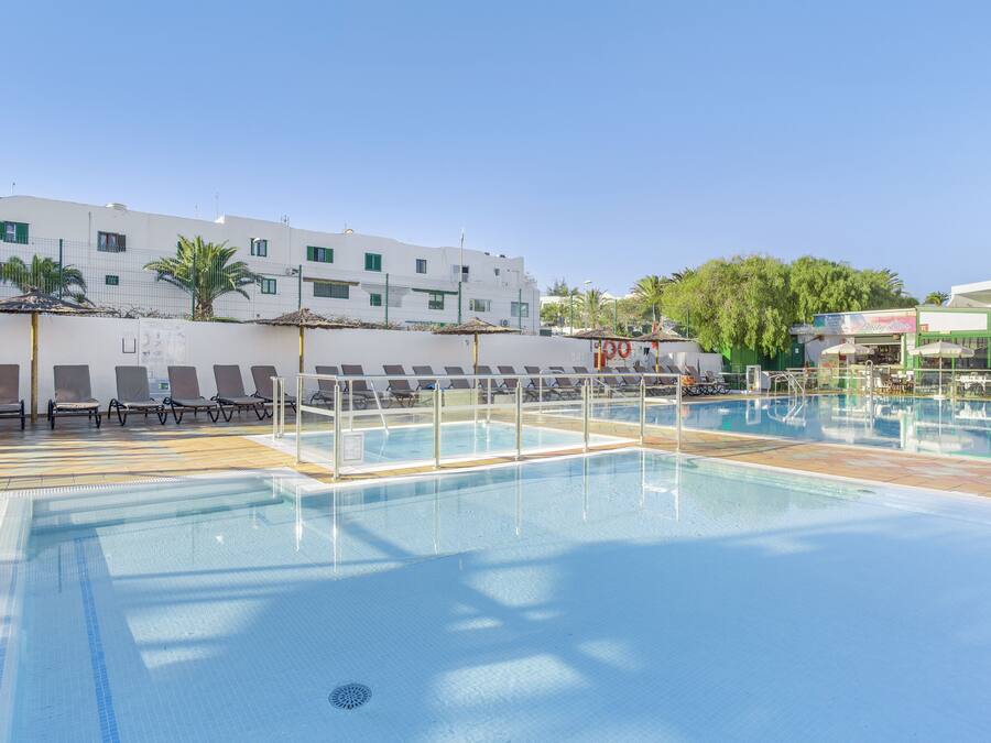 A swimming pool with lounge chairs and umbrellas, a white building with multiple windows, and palm trees in the background.