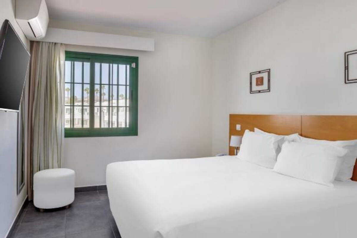 A hotel room with a large bed, a small ottoman, a window with curtains, and a wall-mounted TV.