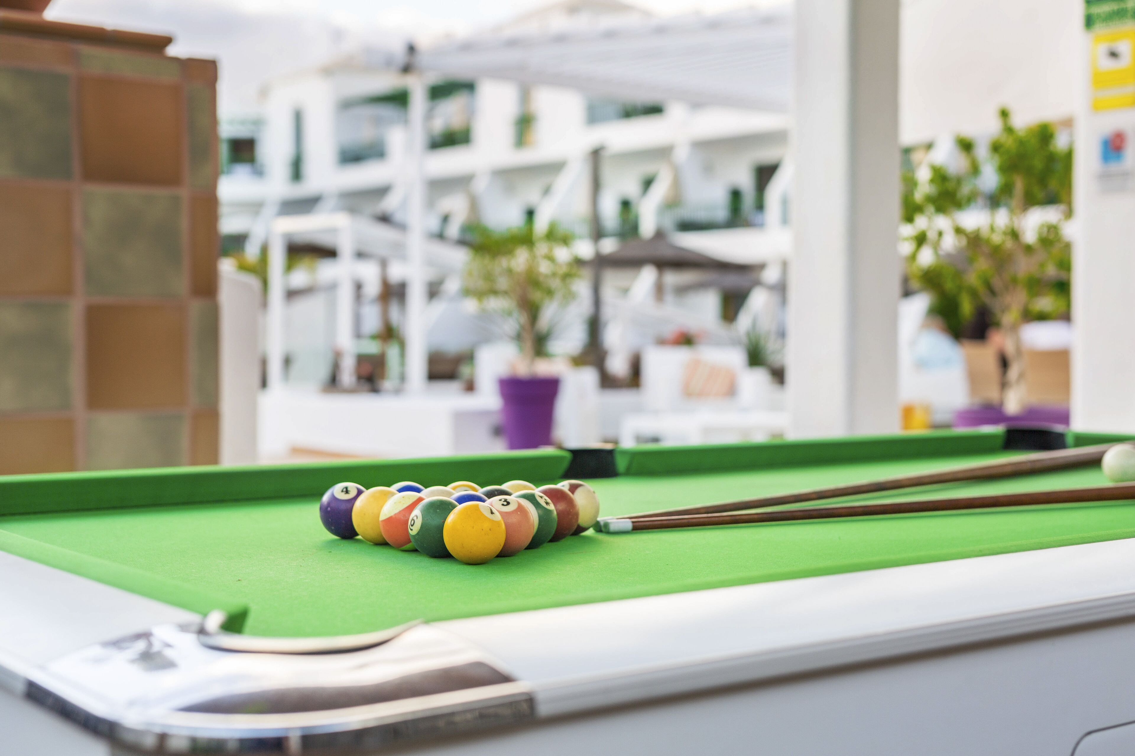 Une table de billard avec des billes et des queues de billard, placée devant un bâtiment moderne doté de balcons et de plantes en pot.