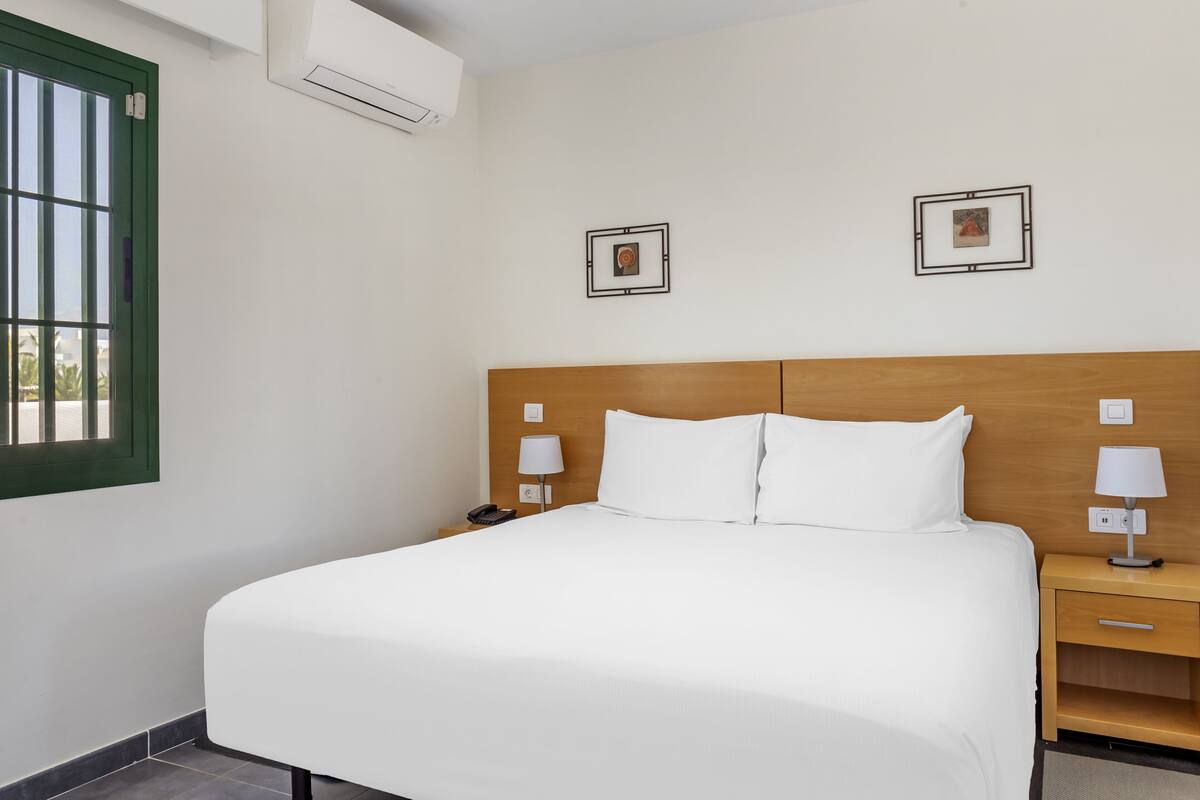 A neatly made bed with white linens, a wooden headboard, two bedside tables with lamps, and two framed pictures on the wall.