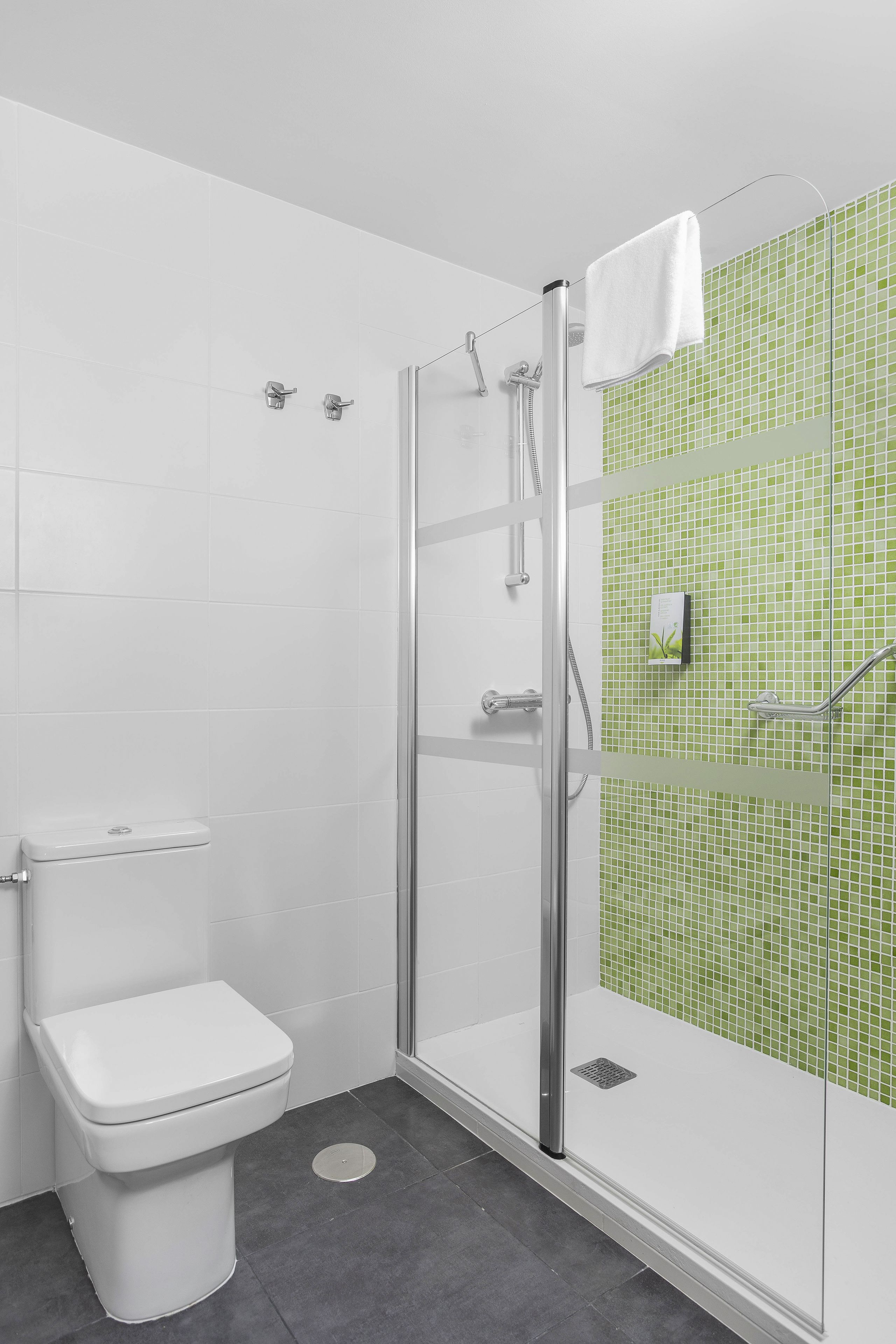 Et moderne badeværelse med en glasafskærmet brusekabine, et hvidt toilet og grønne mosaikfliser.