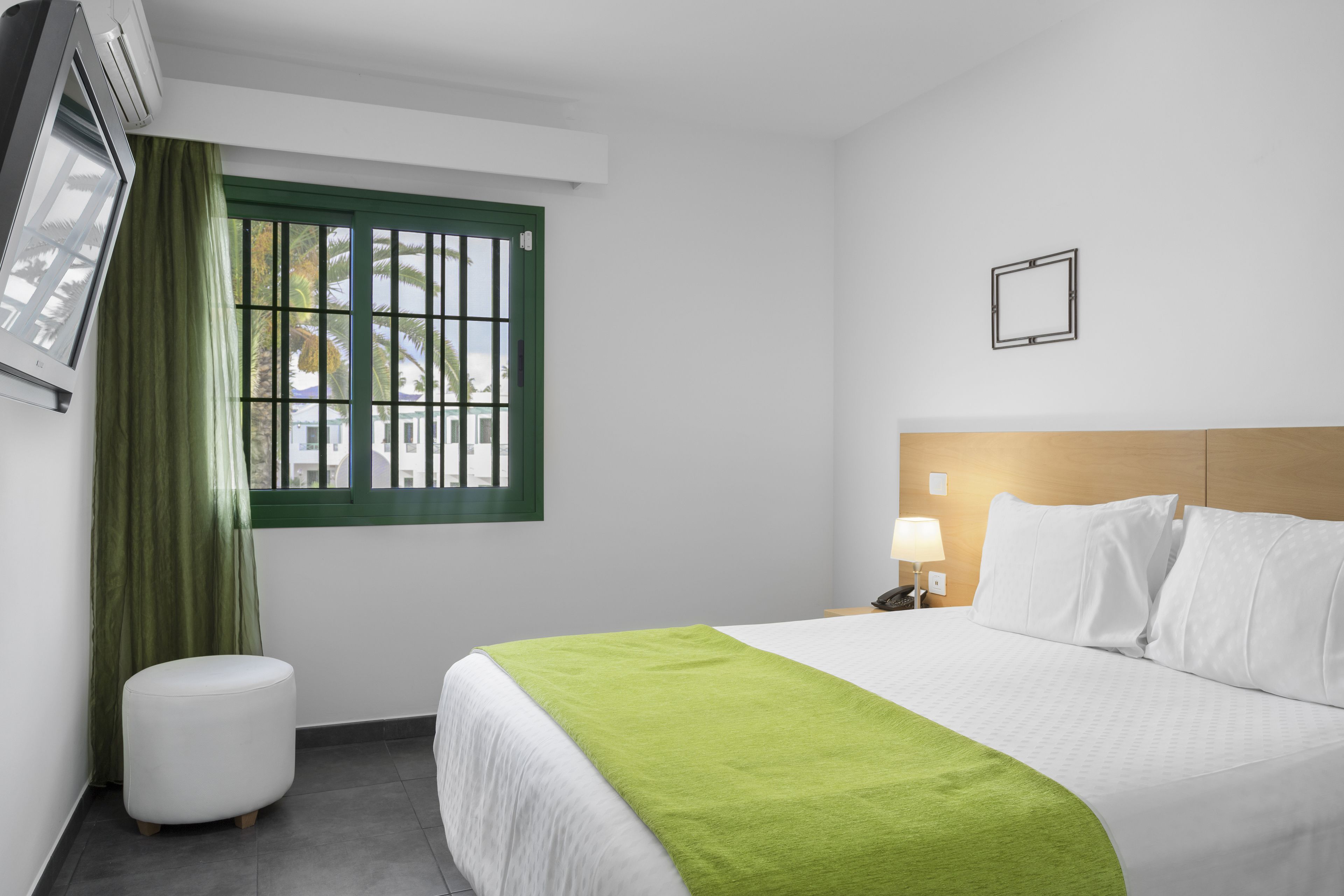 Ein Hotelzimmer mit einem Bett, einem grünen Vorhang, einem weißen Hocker, einem Nachttisch mit einer Lampe und einem Fenster mit Gitterstäben.