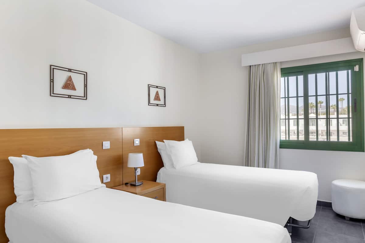 A hotel room with two beds, a wooden headboard, a nightstand with a lamp, a small round table, and a window with curtains.