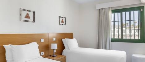 A hotel room with two beds, a wooden headboard, a nightstand with a lamp, a small round table, and a window with curtains.