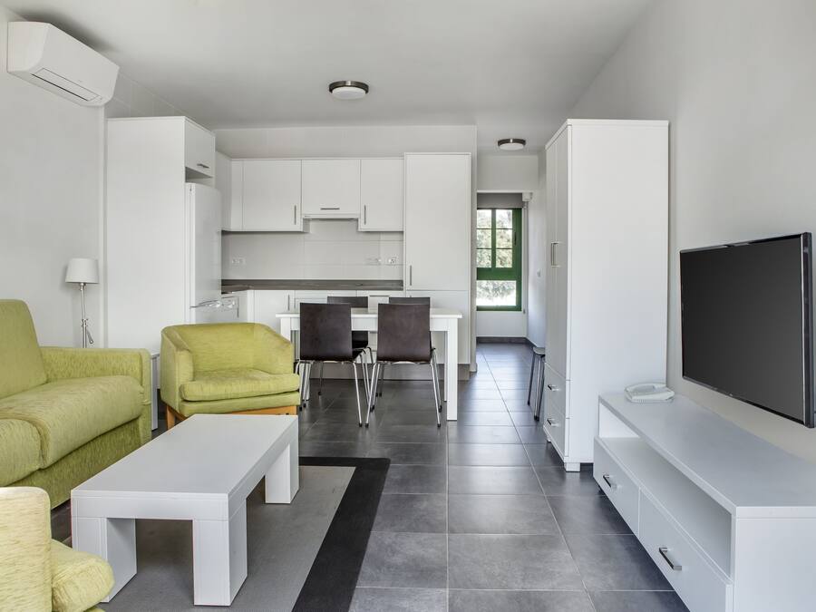 Una sala de estar moderna con sofá, sillas, una mesa de centro, televisor y una cocina al fondo.