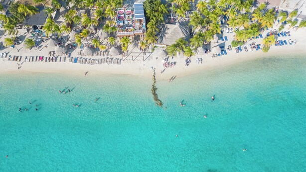 Vista aérea de una playa con aguas turquesas cristalinas, palmeras y una zona de resortes con edificios y sombrillas.