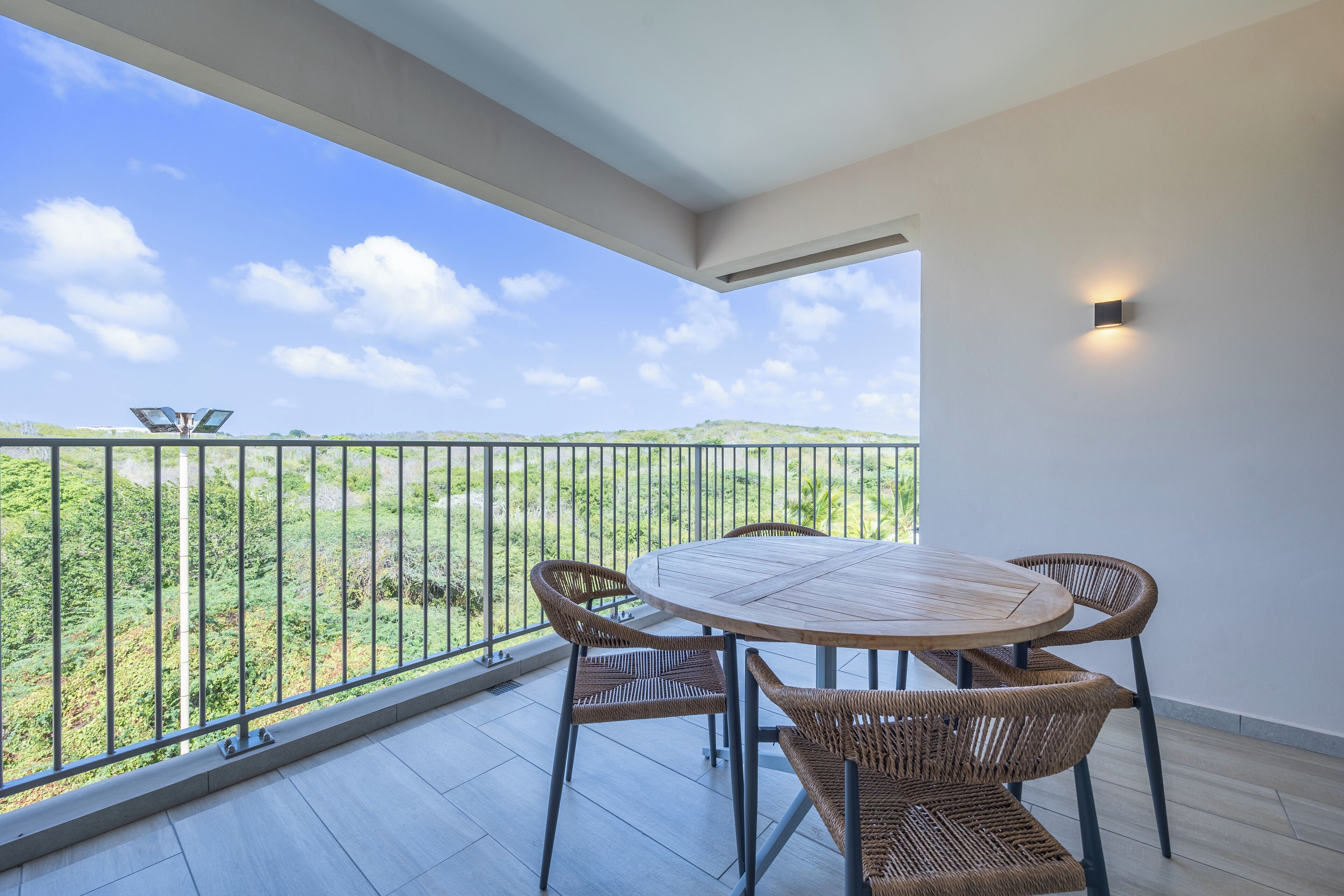 A balcony with a round table and four wicker chairs, overlooking a green landscape with a clear sky.