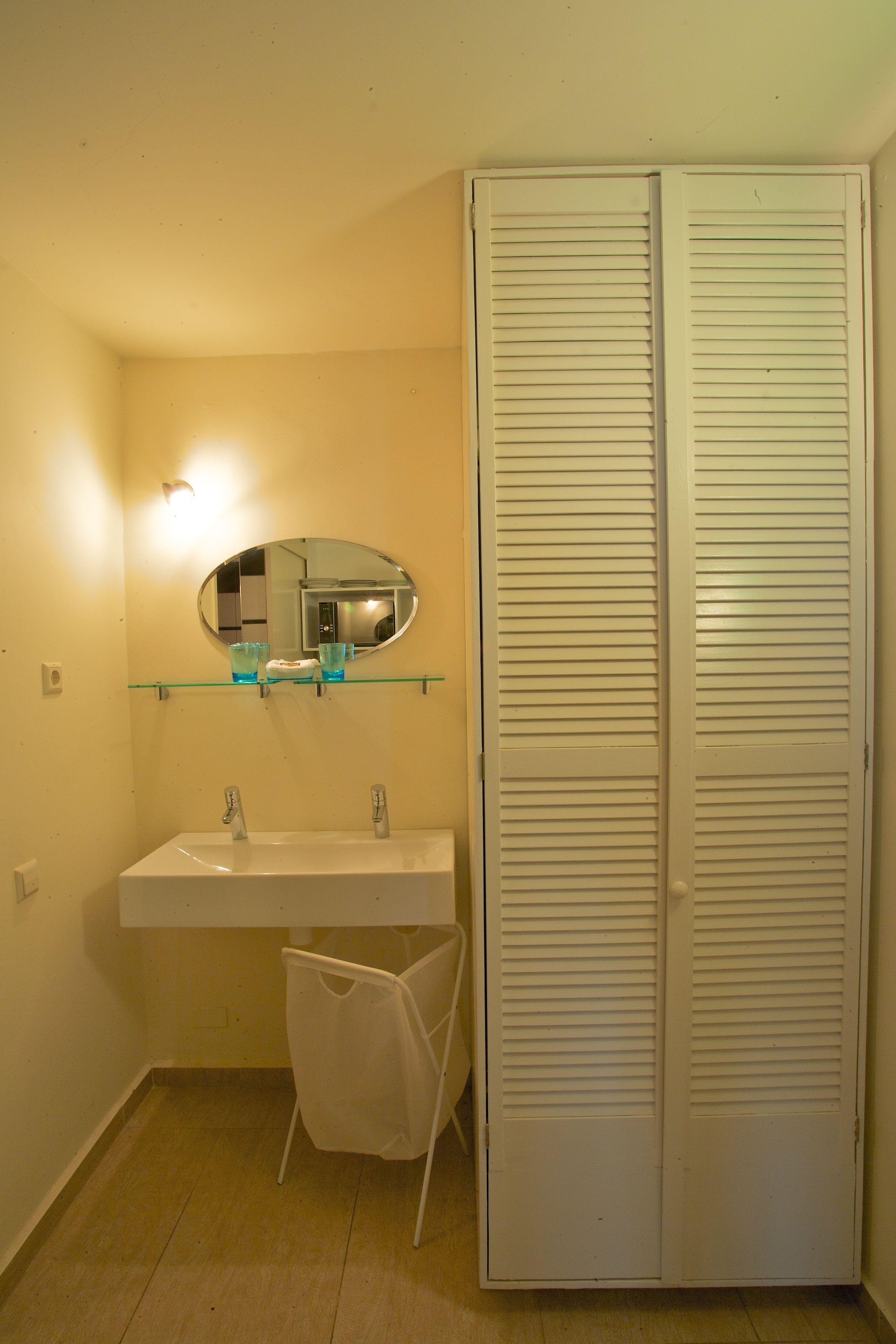 Une salle de bain avec un lavabo blanc, un miroir et une étagère en verre au-dessus. On y trouve également une chaise blanche et un meuble de rangement blanc avec des portes à persiennes.