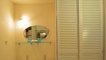 Un baño con un lavabo blanco, un espejo y un estante de vidrio encima. Hay una silla blanca y un mueble de almacenamiento blanco con puertas empañadas.