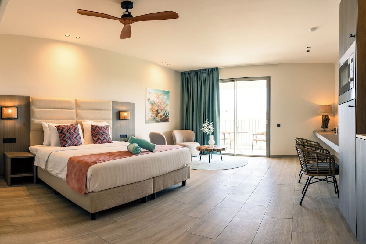Un dormitorio de hotel moderno con una cama grande, un ventilador de techo, un rincón de descanso con sillas y vistas a través de un ventanal.