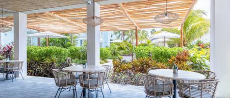 An outdoor patio area with round tables and wicker chairs, surrounded by lush greenery and palm trees.