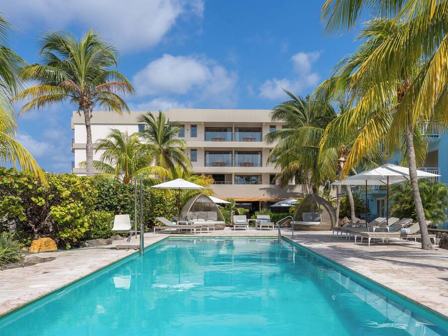 A swimming pool with lounge chairs and umbrellas, surrounded by palm trees and a building with balconies.