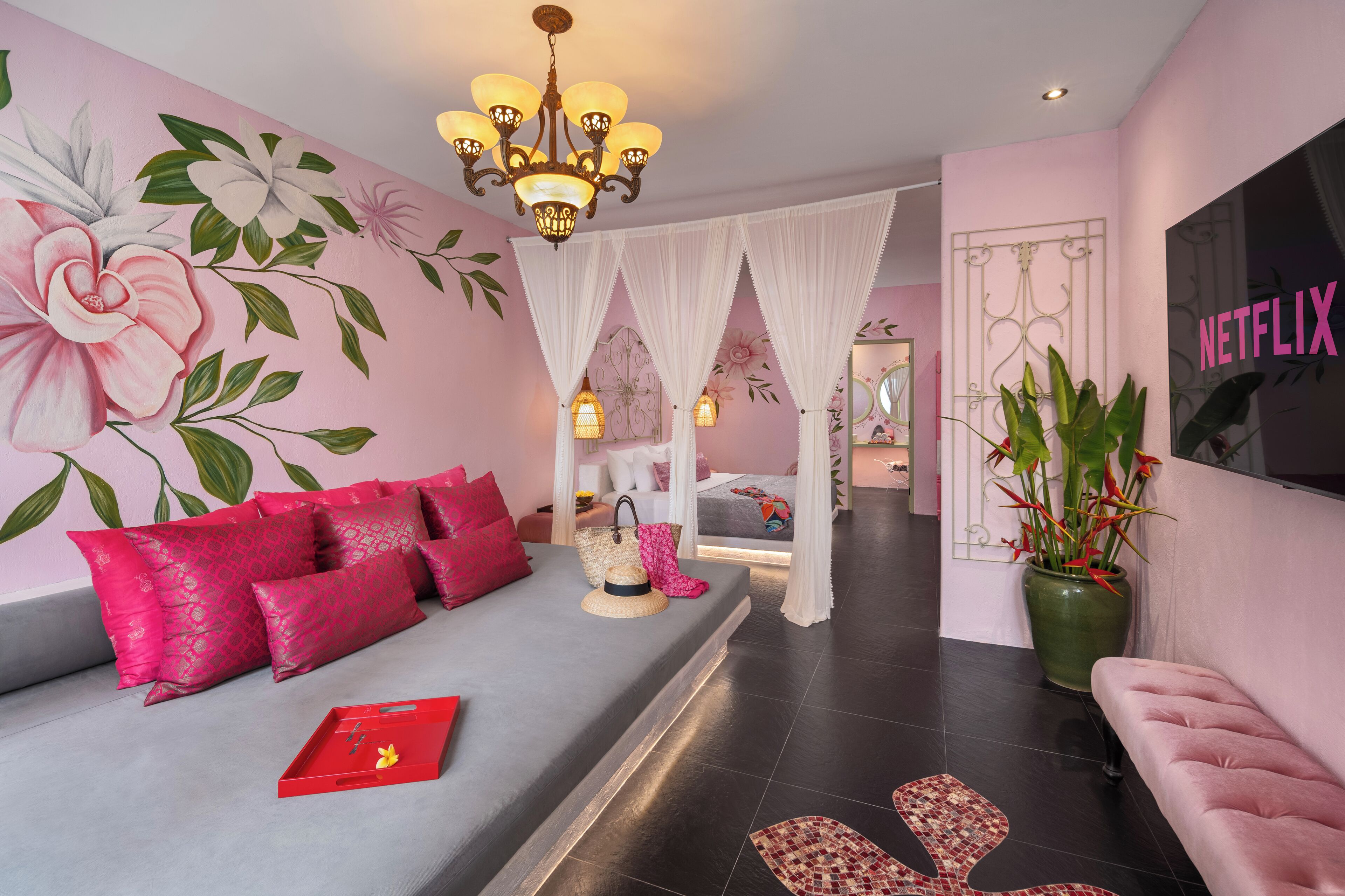 A modern hotel room with a grey sofa, pink pillows, a red tray, a large potted plant, and a Netflix sign on the wall.