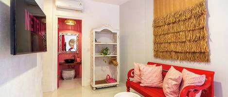 A room with a red bench, a white shelf, a wall-mounted TV, and a red door leading to a bathroom.
