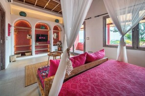 A bedroom with a large bed, pink bedding, a TV on a shelf, and a view of the outdoors.