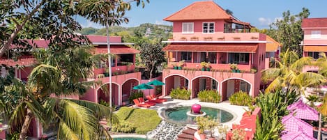 Un resort con piscina central, varias construcciones de color rosa y exuberante vegetación tropical.