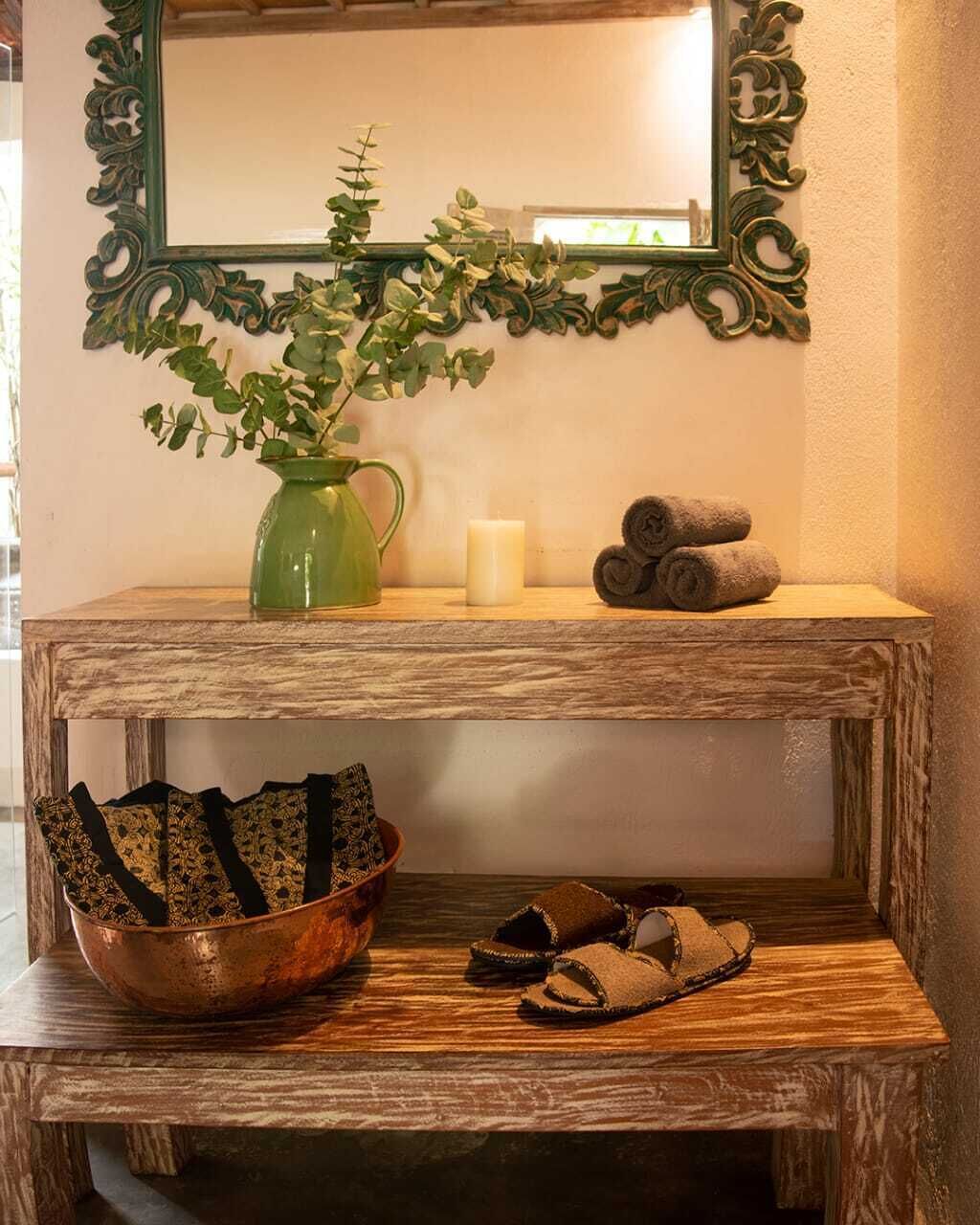 A wooden console table with a mirror, a green vase with plants, a candle, and rolled towels.