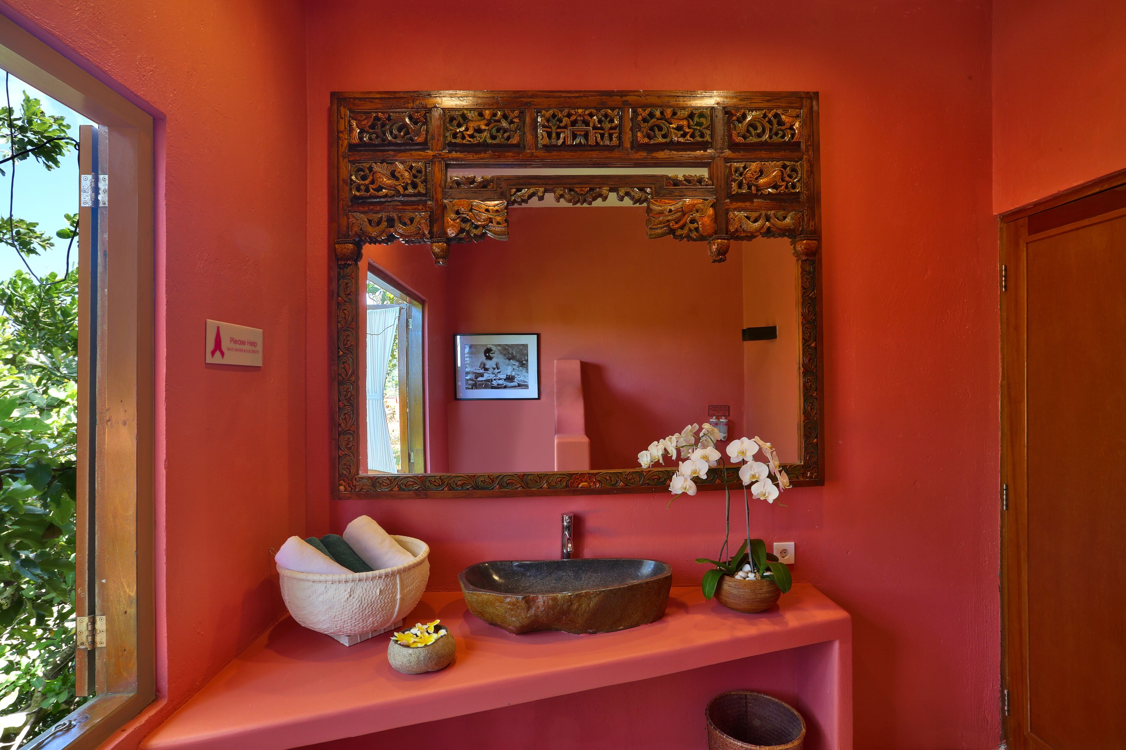 A bathroom with a pink wall, a wooden-framed mirror, a stone sink, a white bowl, and a small plant.