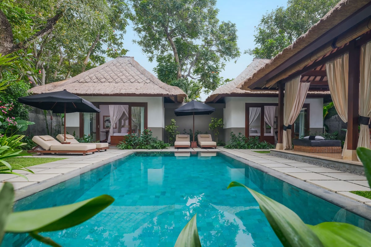 A pool area with lounge chairs and umbrellas, surrounded by lush greenery and thatched-roof structures.