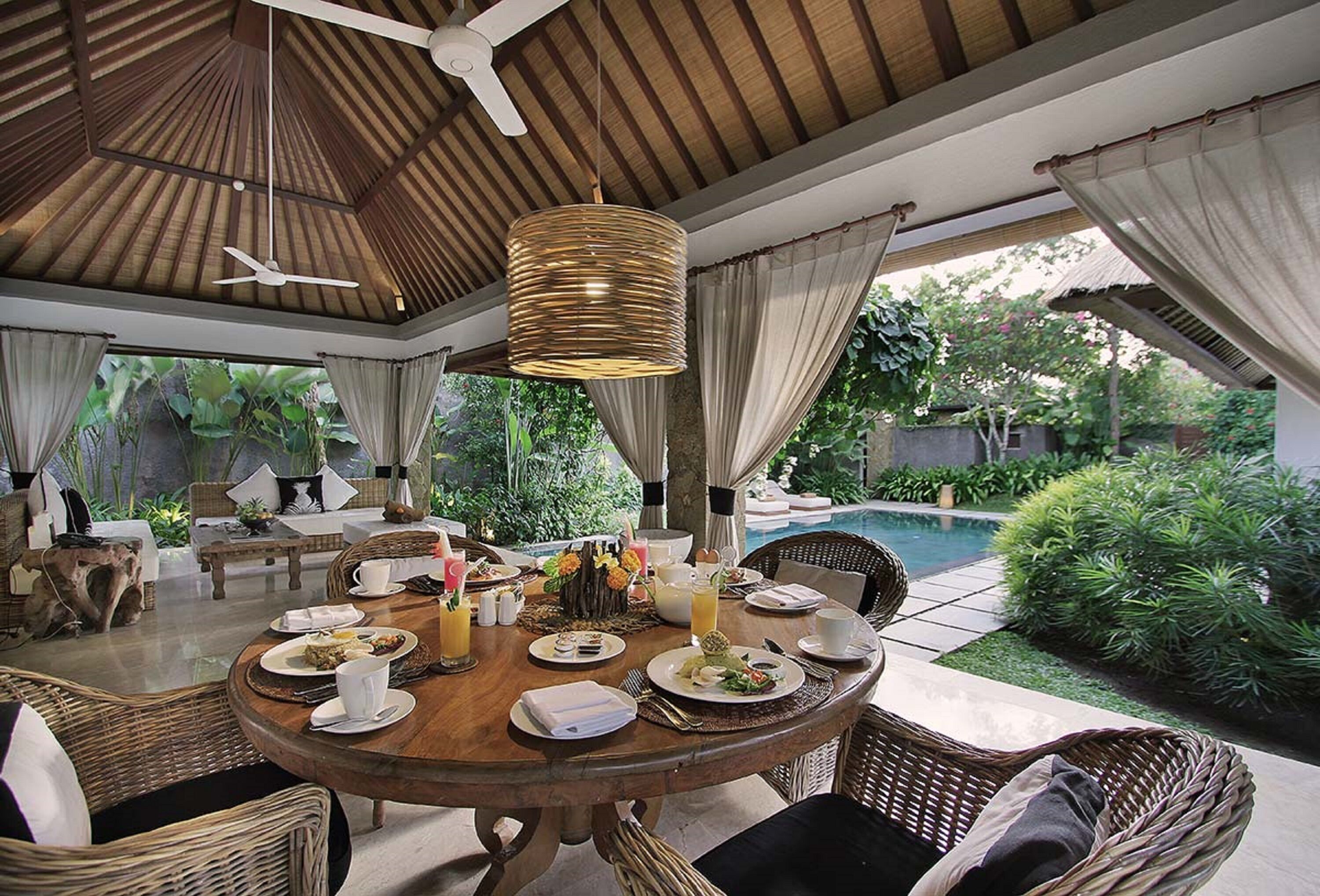 An outdoor dining area with wicker furniture, a round wooden table, and a hanging light fixture.
