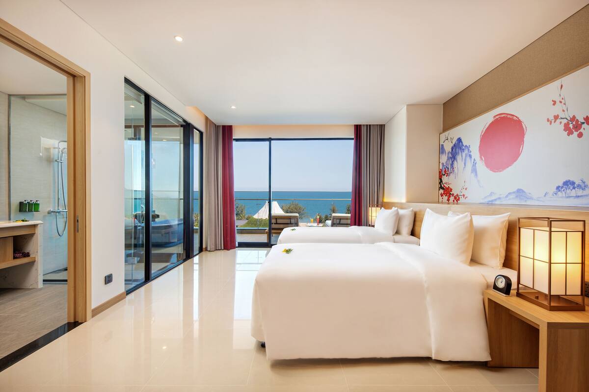Een hotelkamer met een groot bed, een nachtkastje met een lamp en uitzicht op zee door een glazen deur.