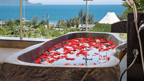 Una bañera llena de agua y pétalos de rosa, con vistas a un paisaje marino y un volcán distante.