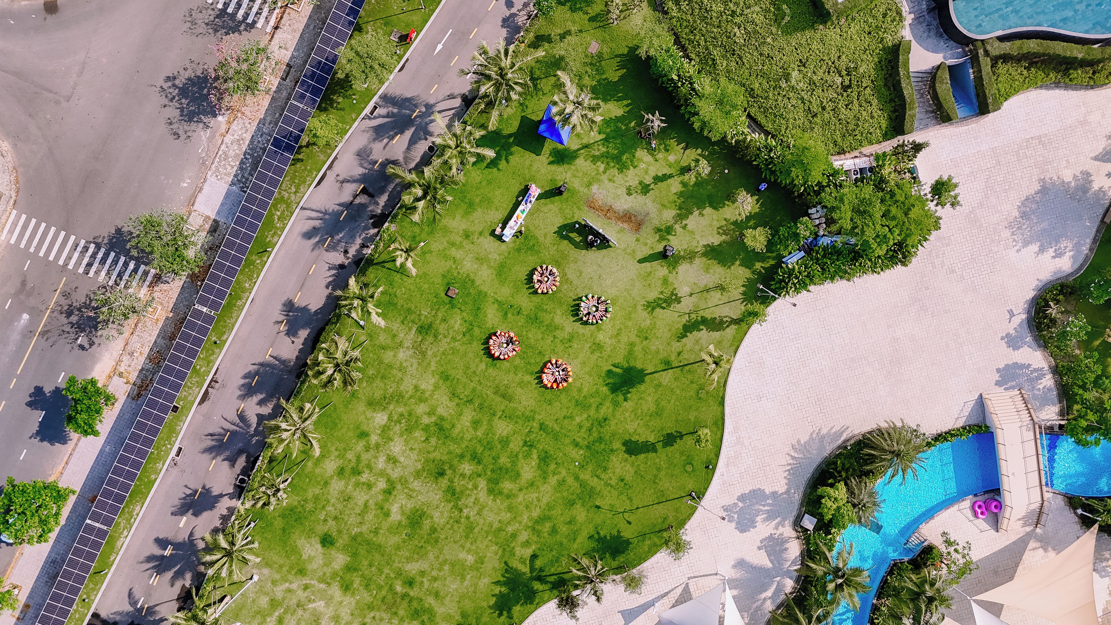 Eine Vogelperspektive zeigt ein Resort mit Schwimmbad, Palmen und Solaranlagen am Straßenrand.