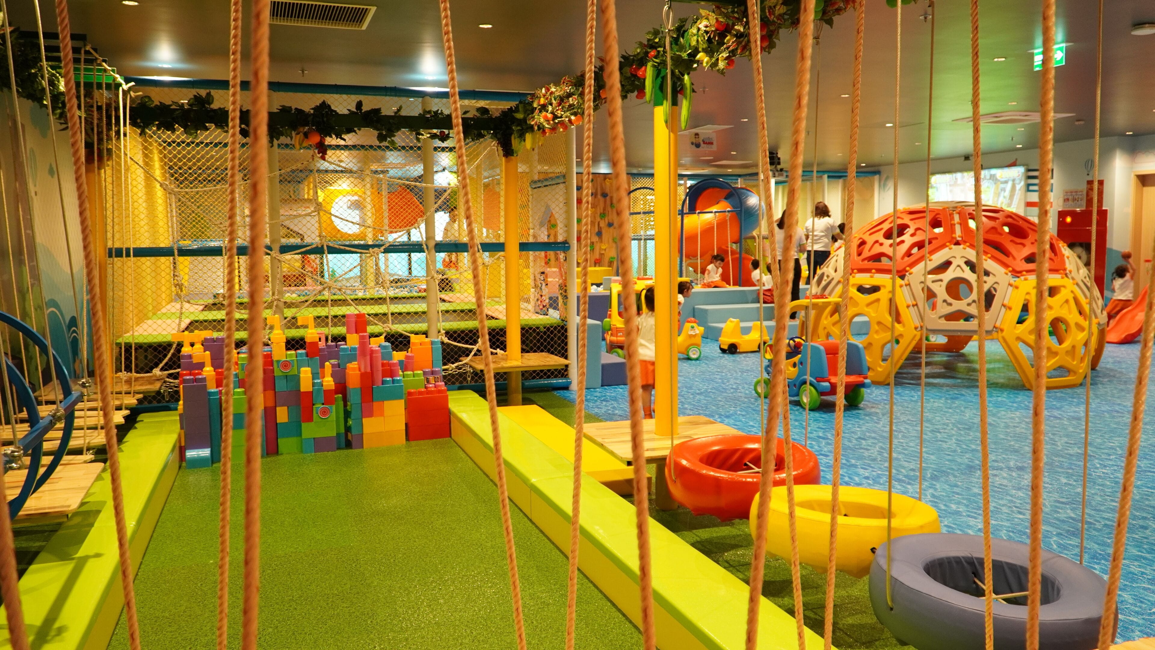 Un espace de jeux intérieur doté de diverses structures ludiques, notamment un filet d’escalade, des revêtements de sol souples et du matériel de jeu coloré.