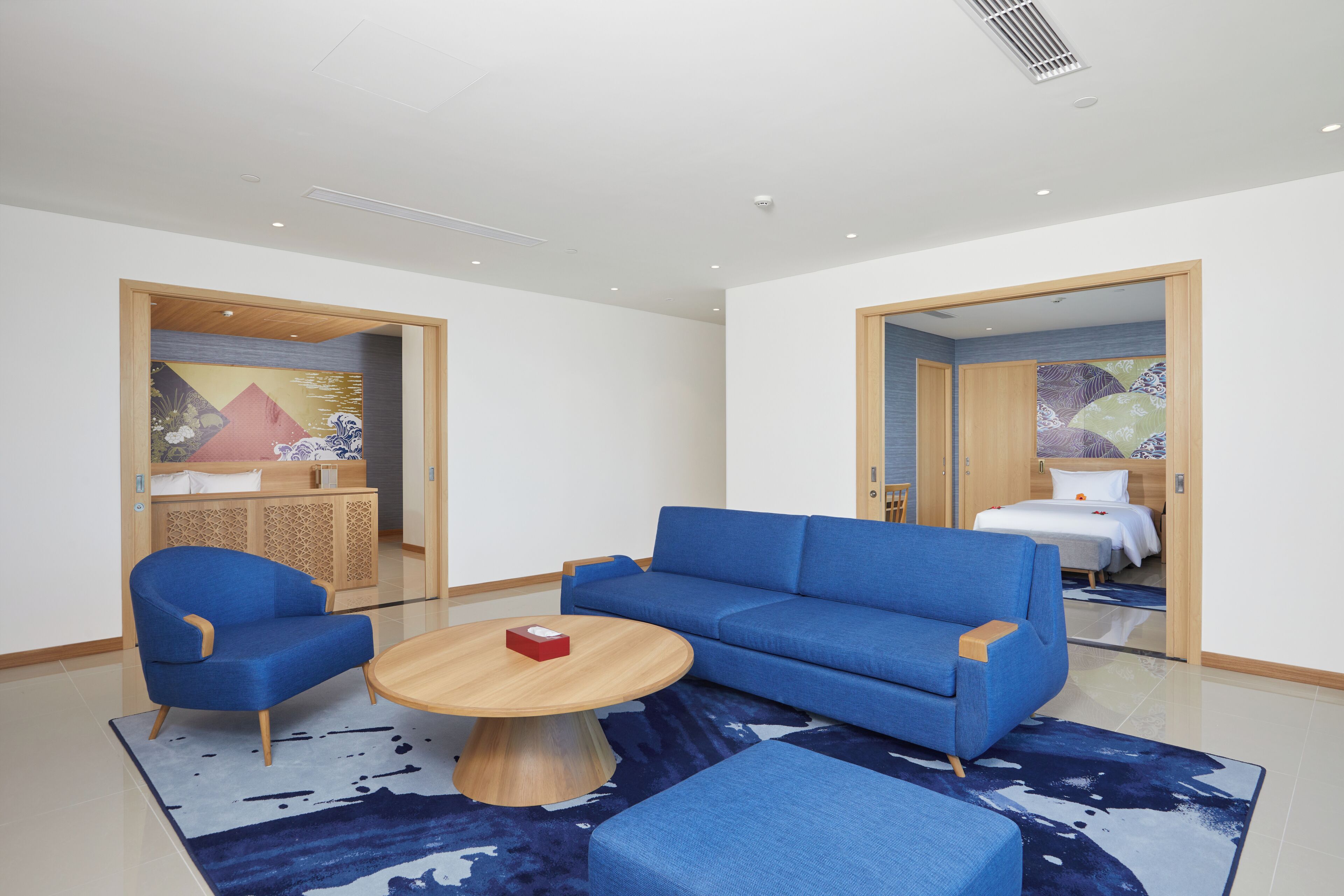 A modern hotel room with a blue sofa, a wooden coffee table, and a blue armchair. There is a bed with white bedding and a wooden headboard in the background.