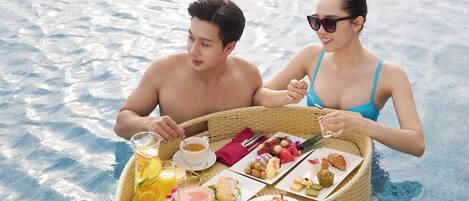 Duas pessoas em uma piscina, com uma bandeja flutuante com comida e bebidas.