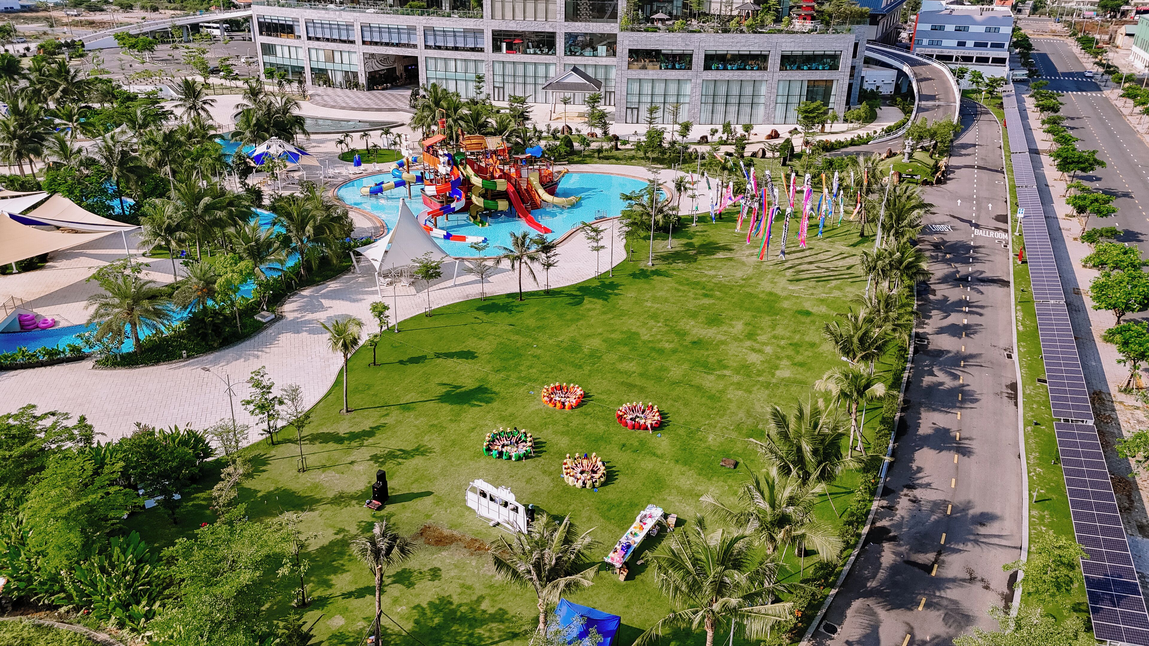 An aerial view of a resort with a large swimming pool, palm trees, and a modern building complex.